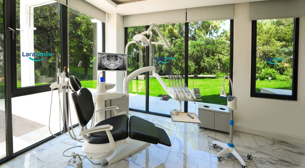 Modern dental treatment room with dental chair, imaging equipment, and large windows overlooking a garden at Lara Smile Dental Clinic in Antalya, Turkey.